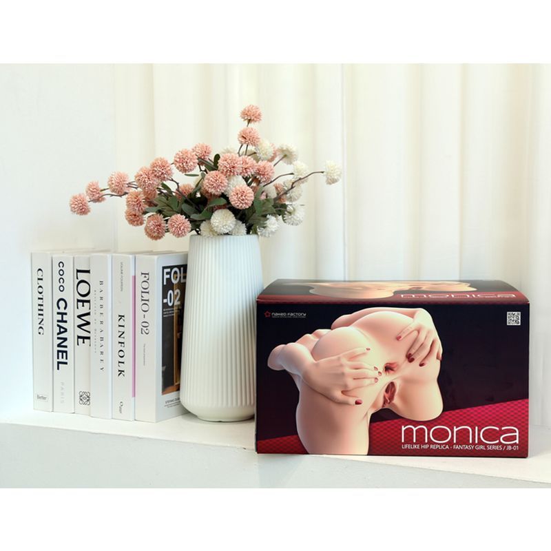 Dual entry torso masturbator Monica box displayed alongside decorative flowers and books.