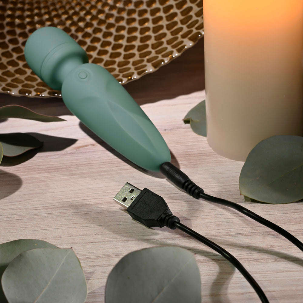 Evolved RUMBLE WAND - Olive 13.4 USB Rechargeable Mini Massage Wand on a table with charging cable and candles.