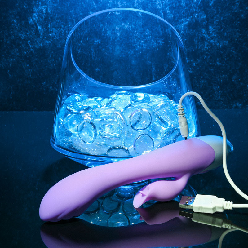 Evolved PLEASING PETAL - Lilac 19.7 cm USB Rechargeable Rabbit Vibrator beside an illuminated glass bowl.