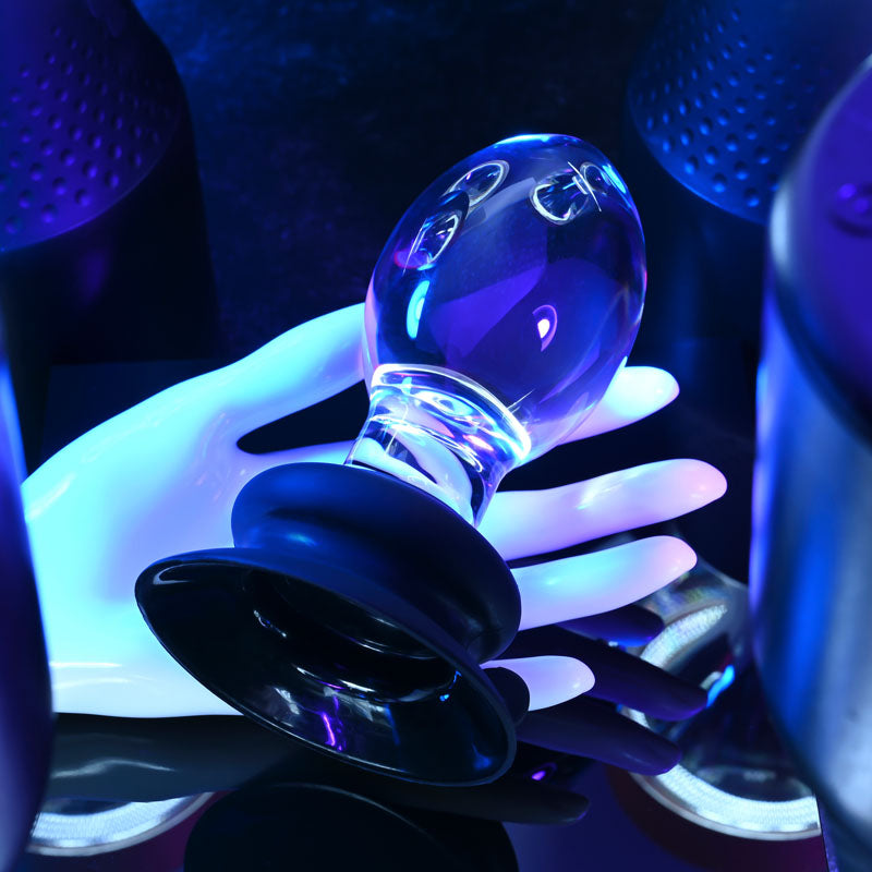 Gender X Crystal Ball -  Glass 13.6 cm Butt Plug with Suction Base placed on a hand in a vibrant blue lit setting.