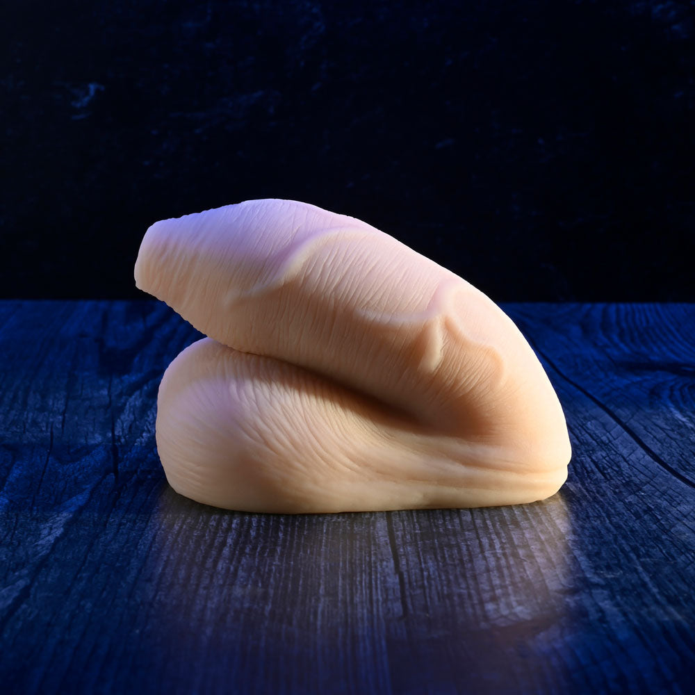 Gender X THE UNCIRCUMCISED PACKER - Light - 11.5 cm Packer displayed on wooden surface with dim lighting.
