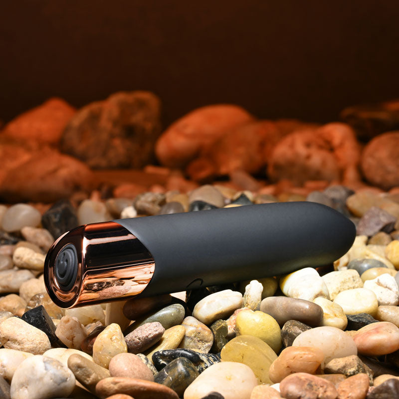 Gender X THE GOLD STANDARD - /Rose Gold 10 cm USB Rechargeable Bullet resting on smooth pebbles, showcasing its sleek design.