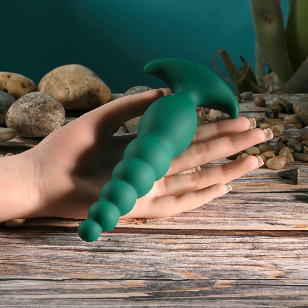 Hand holding a Gender X THE RATTLER USB rechargeable vibrating anal plug on a table with pebbles.