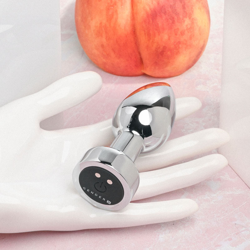 Gender X ROCKIN' METAL PLUG - Metallic 9.3 cm USB Rechargeable Butt Plug on a display with a peach in the background.