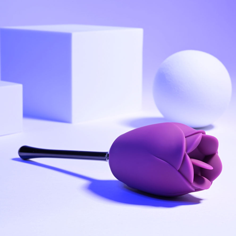 Playboy Pleasure PETAL - USB Rechargeable Flicking Stimulator resting on a surface surrounded by geometric shapes and a ball.