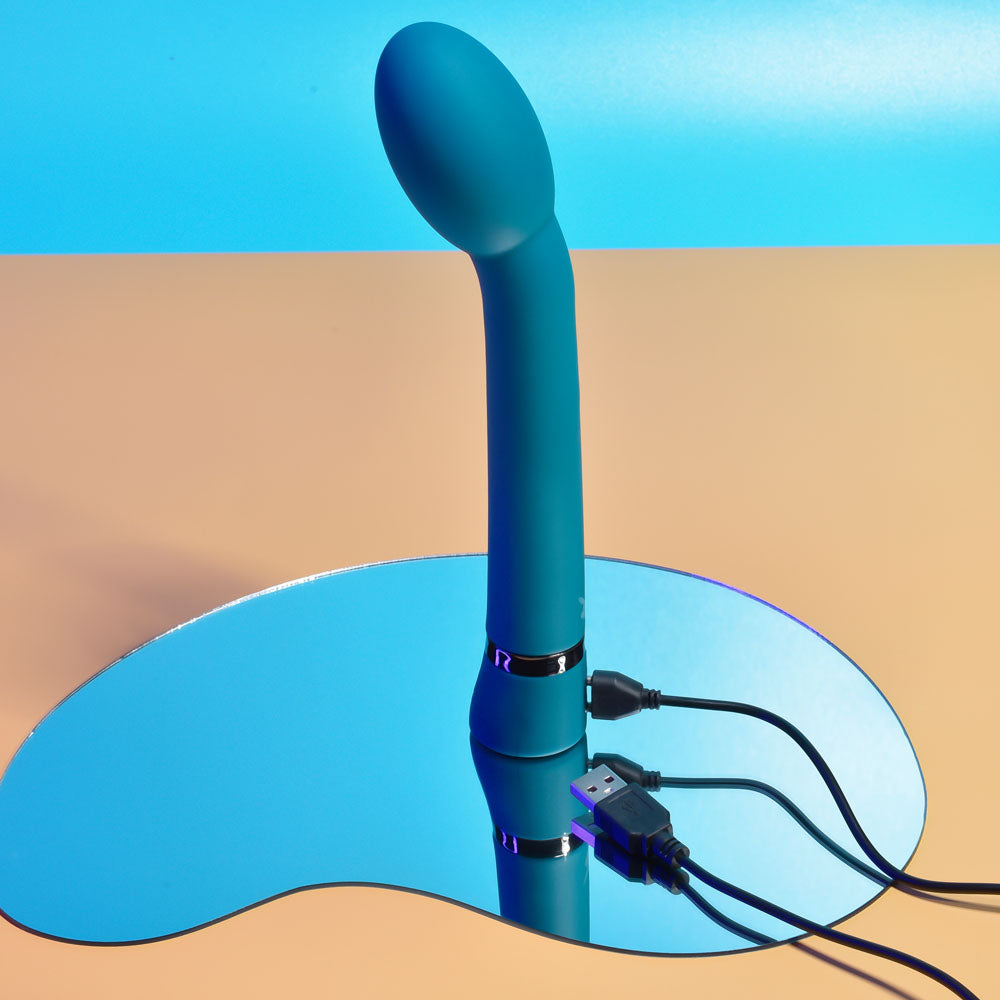 Playboy Pleasure ON THE SPOT - 21.6 cm USB Rechargeable G-Spot Vibrator on a mirror surface with charging cables.