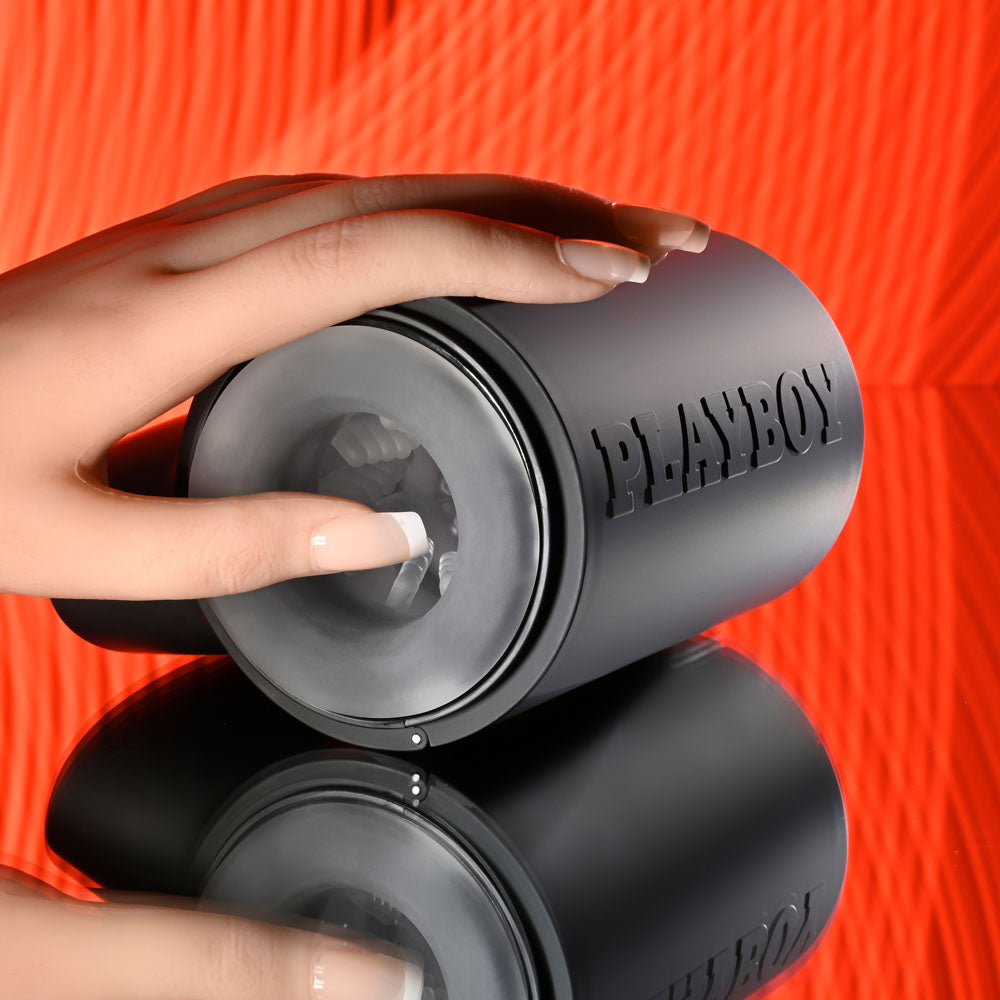 Playboy Pleasure COME ALONG - USB Rechargeable Vibrating Stroker with a hand gripping the canister against a reflective surface.