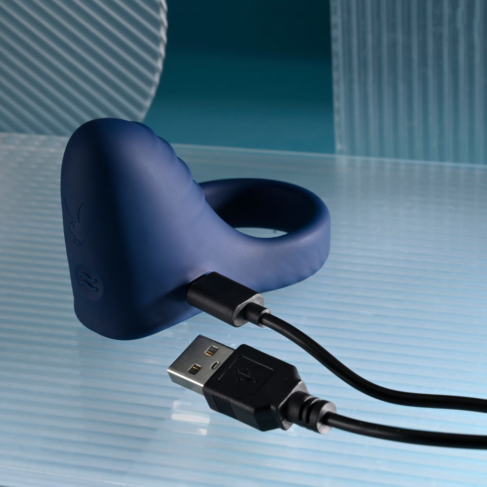 Playboy Pleasure PLEASURE POINT - USB Rechargeable Vibrating Cock Ring with charging cable on a sleek surface.