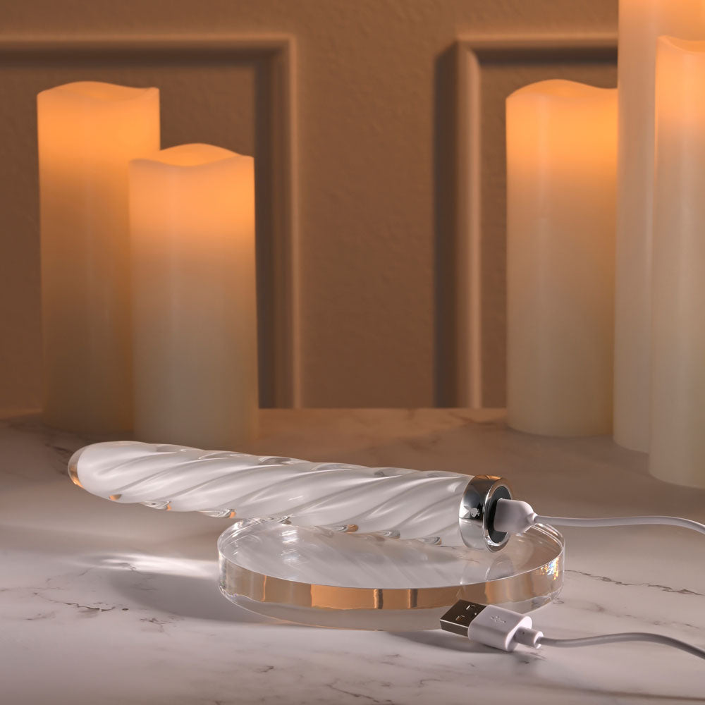 Playboy Pleasure IN A TWIST - Glass 17.5 cm USB Rechargeable Vibrator on a charging base with soft candlelight.