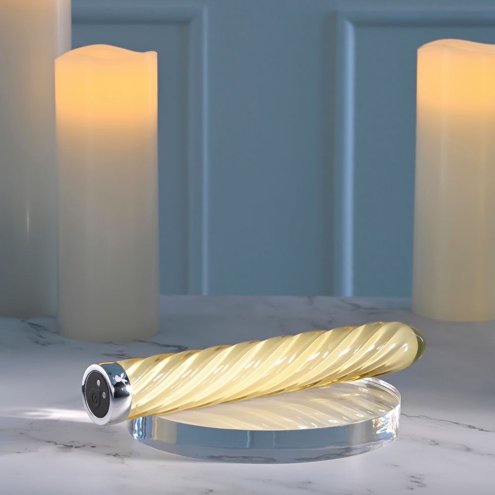 USB rechargeable glass vibrator with a chic twisted design, placed on a reflective surface beside soft glowing candles.